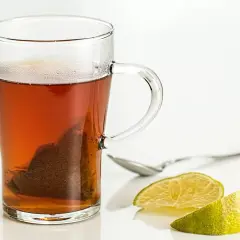 Tè caldo in tazza di vetro con bustina, limone e cucchiaino. Scopri i tè al limone benefici per il tuo benessere.