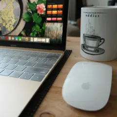 Tazza con scritta "Coffee addict", laptop e mouse su un tavolo di legno. La dipendenza da caffeina è evidente.