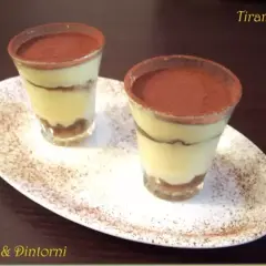 Due tiramisù monoporzione con savoiardi, presentati in bicchieri e spolverati di cacao, su un piatto bianco.