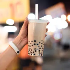 Un bicchiere di bubble tea con perle nere, tenuto da una mano con un braccialetto bianco. Un sorso di freschezza che ricorda le sue origini asiatiche.