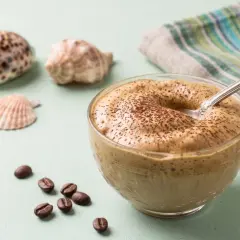 Un cremoso dalgona coffee, spolverato di cacao, accanto a chicchi di caffè e conchiglie.
