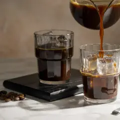 Versando caffè freddi in bicchieri con ghiaccio. Chicchi di caffè tostati e un cucchiaino completano la scena.