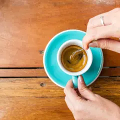 Mani che mescolano un caffè, nonostante si dica che il caffè è cancerogeno, la tazzina turchese su un tavolo di legno è invitante.