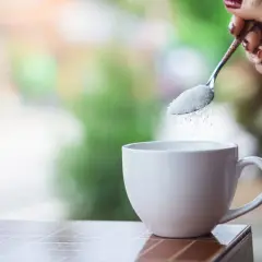 Mano con cucchiaino che versa zucchero in una tazza bianca, pronta per il caffè.
