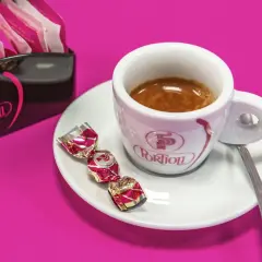 Una tazzina di crema al caffè espresso, con tre caramelle incartate e un porta-bustine Portioli su sfondo fucsia.