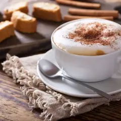 Un invitante cappuccino caldo, spolverato di cacao, accanto a biscotti croccanti e cannella su un tavolo di legno.