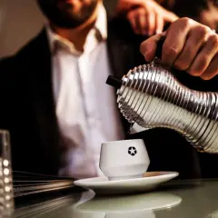 Un uomo in giacca e camicia versa caffè da un bricco argentato in una tazzina bianca. La macinatura caffè più fine garantisce un aroma intenso.
