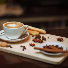 Un cappuccino macchiato con schiuma a forma di cuore, biscotti, chicchi di caffè e spezie su un tagliere di legno.