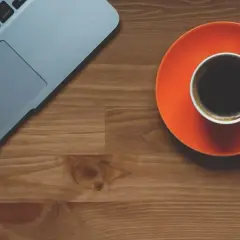 Tazza di caffè nero su piattino arancione accanto a un laptop. Scopri quando bere il caffè per dimagrire.