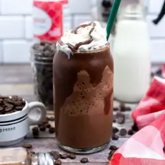 Un delizioso Frappuccino Java Chip, con panna montata e sciroppo al cioccolato. Sullo sfondo, chicchi di caffè, gocce di cioccolato e latte, tutti ingredienti per prepararlo.