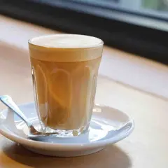 Un bicchiere di cafe latte con schiuma cremosa, servito su un piattino con un cucchiaino, su un tavolo di legno.