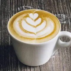 Un flat latte con schiuma artistica a forma di cuore e petali, servito in una tazza bianca su un tavolo di legno scuro.