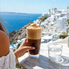 Una mano tiene un bicchiere di caffè greco freddo con schiuma abbondante, con vista su un villaggio bianco sul mare.