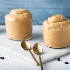 Due bicchieri di soffice crema al caffè senza panna, preparata con caffè moka, su un tagliere di marmo con chicchi di caffè e cucchiaini dorati.