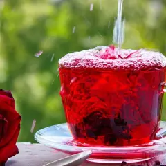 Tè rosso vibrante versato in una tazza, con una rosa rossa accanto. Scopri le proprietà e le controindicazioni dell'ibisco.