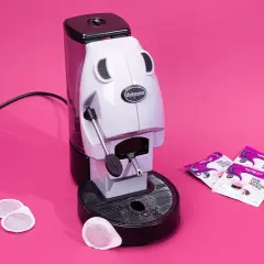 Didiesse Frog espresso machine with coffee pods and Lavazza Gran Espresso Intenso packets on a pink background.