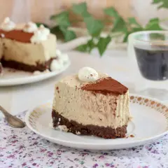 Una fetta di cheesecake al caffè senza gelatina, con base croccante al cacao e decorata con panna e cacao in polvere.