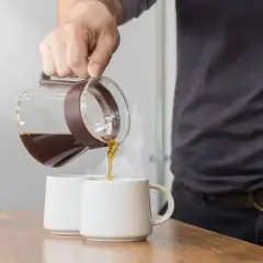 Un uomo versa caffè americano con latte in due tazze bianche su un tavolo di legno.