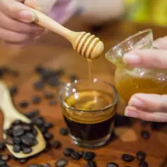 Gocce dorate di miele scendono su un caffè espresso. Chicchi di caffè tostati e un cucchiaino di legno completano la scena.