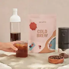 Un bicchiere di caffè freddo in bottiglia, pronto per essere gustato. Accanto, una confezione di cold brew e un macinacaffè.