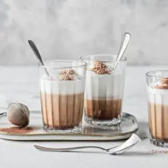 Tre bicchieri di cappuccino con cioccolato, guarniti con panna e cacao, su un vassoio con colino.
