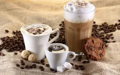 Tre tazze di caffè, una con panna e cacao, un'altra con schiuma e un bicchiere alto di latte cappuccino, accompagnate da biscotti e chicchi di caffè.