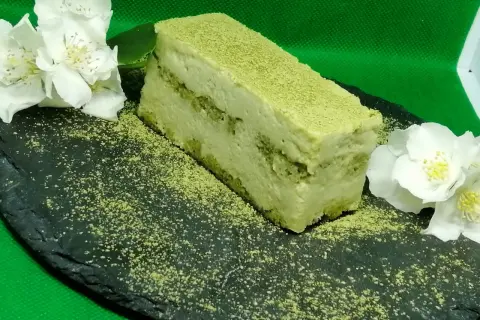 Un delizioso matcha tiramisù, spolverato con polvere verde, adagiato su una pietra scura con fiori bianchi.