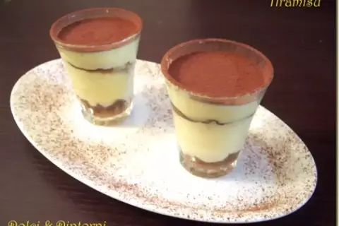 Due tiramisù monoporzione con savoiardi, presentati in bicchieri e spolverati di cacao, su un piatto bianco.