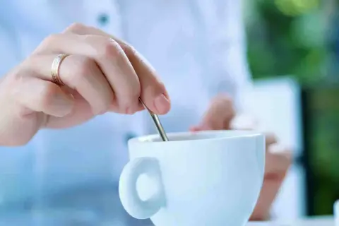 Mano con anello mescola il caffè in una tazza bianca. La scelta è sempre la stessa: caffè con o senza zucchero.
