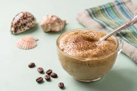 Un cremoso dalgona coffee, spolverato di cacao, accanto a chicchi di caffè e conchiglie.