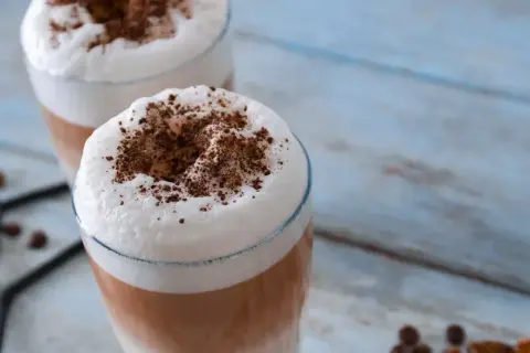 Due bicchieri di latte macchiato freddo con schiuma soffice e cacao in polvere, su un tavolo di legno azzurro.