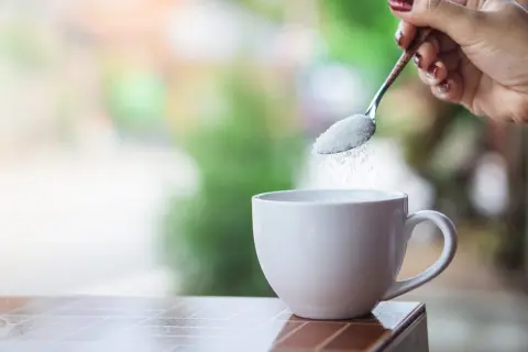 Mano con cucchiaino che versa zucchero in una tazza bianca, pronta per il caffè.