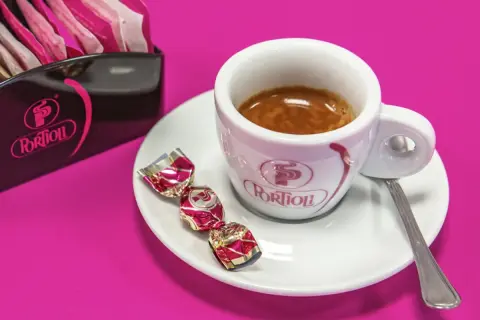 Una tazzina di crema al caffè espresso, con tre caramelle incartate e un porta-bustine Portioli su sfondo fucsia.