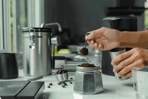 Mano aggiunge caffè macinato a una moka, per il suo primo utilizzo. Caffè in grani e contenitori in acciaio completano la scena.