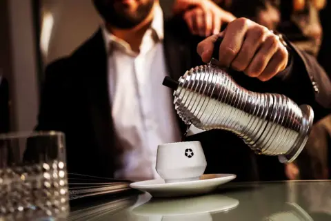 Un uomo in giacca e camicia versa caffè da un bricco argentato in una tazzina bianca. La macinatura caffè più fine garantisce un aroma intenso.