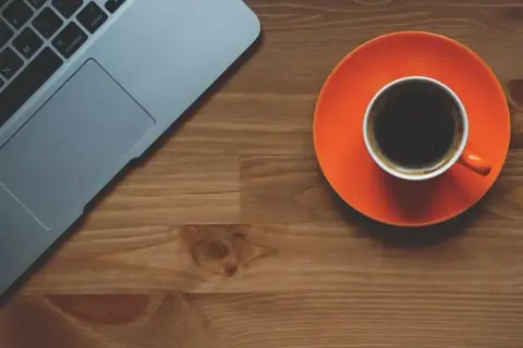 Tazza di caffè nero su piattino arancione accanto a un laptop. Scopri quando bere il caffè per dimagrire.