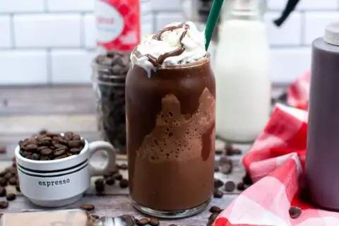 Un delizioso Frappuccino Java Chip, con panna montata e sciroppo al cioccolato. Sullo sfondo, chicchi di caffè, gocce di cioccolato e latte, tutti ingredienti per prepararlo.