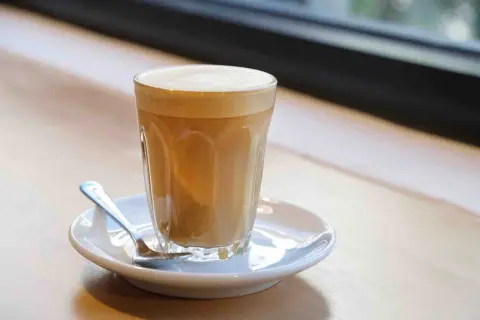 Un bicchiere di cafe latte con schiuma cremosa, servito su un piattino con un cucchiaino, su un tavolo di legno.