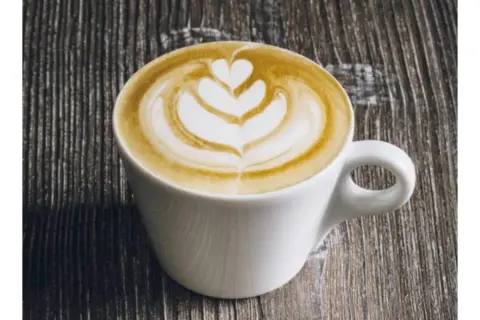 Un flat latte con schiuma artistica a forma di cuore e petali, servito in una tazza bianca su un tavolo di legno scuro.