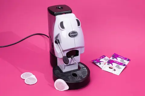 Didiesse Frog espresso machine with coffee pods and Lavazza Gran Espresso Intenso packets on a pink background.