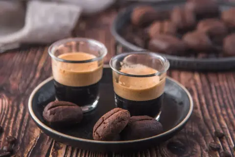 Due bicchierini di espresso con crema e biscotti al caffè moka a forma di chicco, su un piatto scuro.