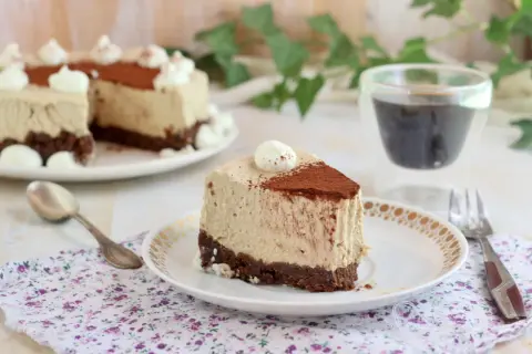 Una fetta di cheesecake al caffè senza gelatina, con base croccante al cacao e decorata con panna e cacao in polvere.