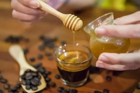 Gocce dorate di miele scendono su un caffè espresso. Chicchi di caffè tostati e un cucchiaino di legno completano la scena.