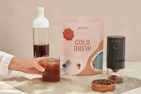 Un bicchiere di caffè freddo in bottiglia, pronto per essere gustato. Accanto, una confezione di cold brew e un macinacaffè.