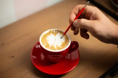 Un barista crea disegni sul cappuccino con uno strumento, mostrando come fare latte art.