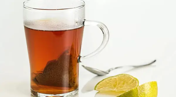 Tè caldo in tazza di vetro con bustina, limone e cucchiaino. Scopri i tè al limone benefici per il tuo benessere.