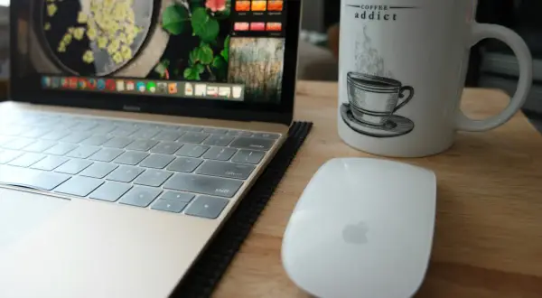 Tazza con scritta "Coffee addict", laptop e mouse su un tavolo di legno. La dipendenza da caffeina è evidente.