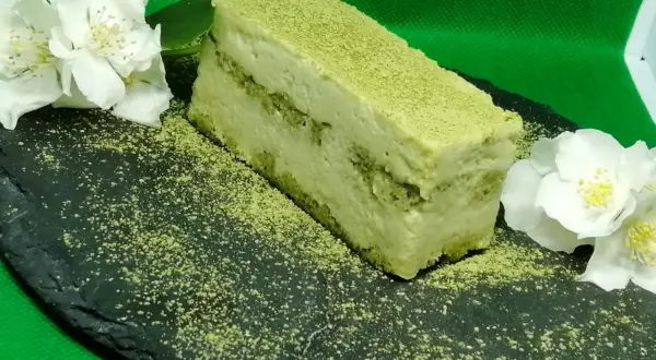 Un delizioso matcha tiramisù, spolverato con polvere verde, adagiato su una pietra scura con fiori bianchi.
