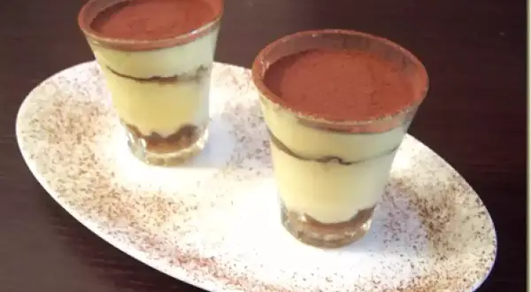 Due tiramisù monoporzione con savoiardi, presentati in bicchieri e spolverati di cacao, su un piatto bianco.