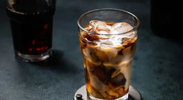 Un bicchiere di **cold brew latte** con ghiaccio, latte e caffè che si mescolano creando un effetto marmorizzato. Sullo sfondo, un altro bicchiere di caffè scuro.